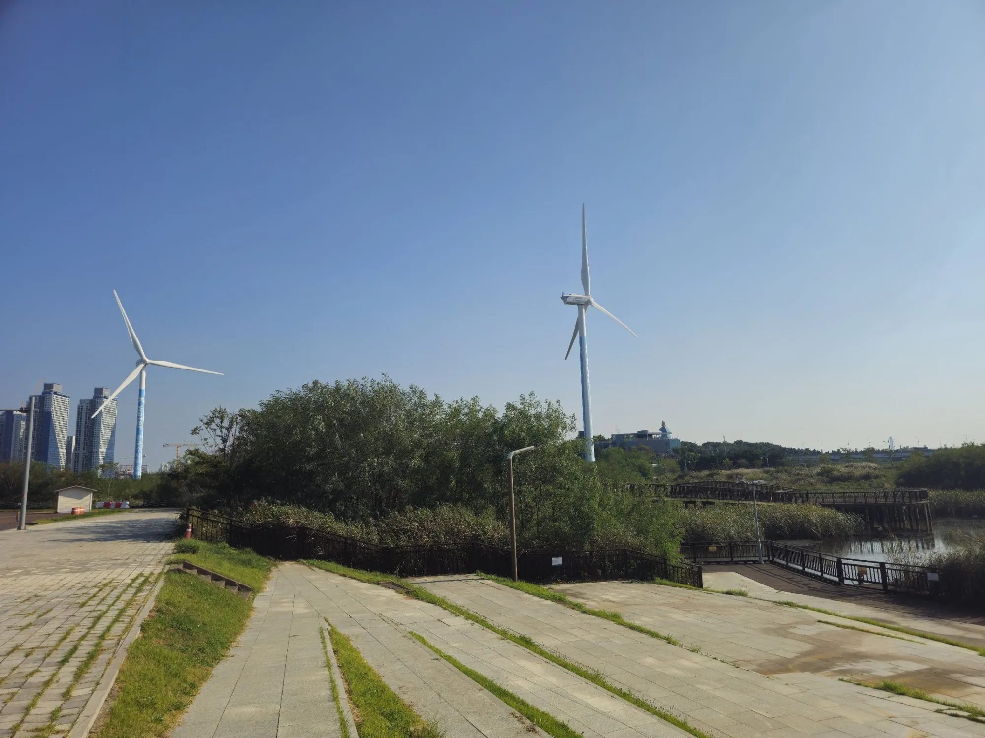 Two wind turbines beside a wooden boardwalk and reed beds at Jeongseojin, with high-rise apartments on the horizon